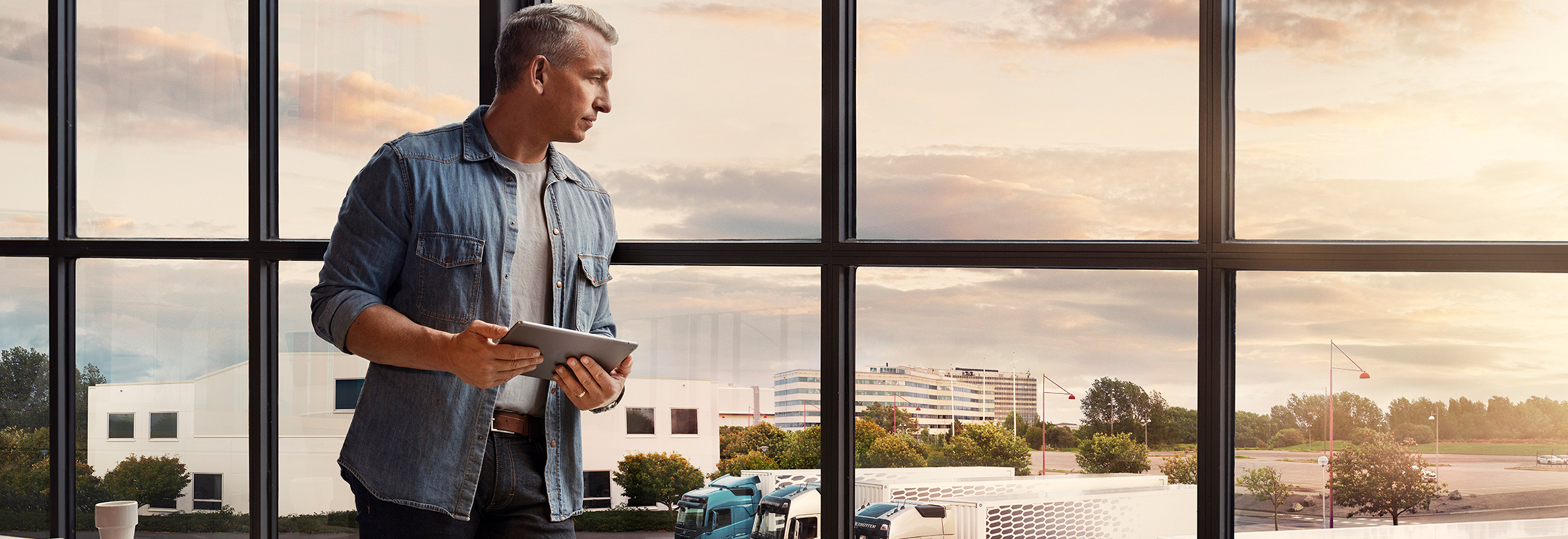 Moški, ki drži tablični računalnik, stoji ob oknu in gleda svoj vozni park